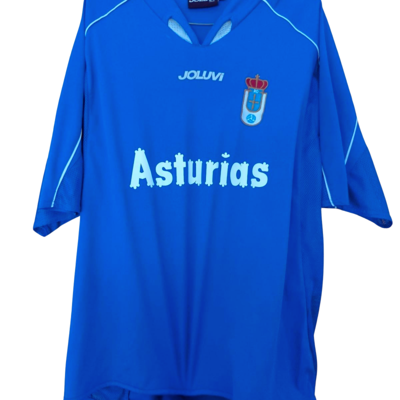 Descripción de la Camiseta del Real Oviedo 2004-2005 (Primera Equipación) Esta es la primera equipación que el Real Oviedo utilizó durante la temporada 2004-2005. Fabricada por la marca asturiana Joluvi, la camiseta es de un vibrante color azul, el tono tradicional del club ovetense. El diseño presenta un cuello en V con una inserción en un tono de azul más claro y un ribete en el mismo tono. Se observan líneas decorativas en azul claro que parten del cuello y recorren la parte superior de los hombros. Las mangas son de color azul, con detalles sutiles en los puños. En el pecho, el logotipo de Joluvi se sitúa debajo del cuello. El escudo del Real Oviedo, que incorpora la corona real, está aplicado en el lado derecho. El patrocinador principal es "Asturias", con la palabra en mayúsculas y un tipo de letra estilizado en color azul claro, que representa el apoyo de la comunidad autónoma al club en un periodo complicado de su historia. Rendimiento del Real Oviedo en la Temporada 2004-2005 La temporada 2004-2005 fue una campaña en la que el Real Oviedo intentaba recuperar posiciones tras haber descendido administrativamente a Tercera División y haber logrado el ascenso a Segunda División B. Segunda División B (Grupo II): El Real Oviedo finalizó en la 5ª posición de su grupo, quedándose a las puertas de la zona de Play-off de ascenso. Copa del Rey: En la Copa del Rey, el equipo alcanzó la segunda ronda de la competición. Jugadores Clave de la Temporada 2004-2005 La plantilla del Real Oviedo en esta temporada estaba formada por una combinación de jugadores locales y algunos refuerzos. Entre los jugadores destacados se encontraban, el delantero Diego Cervero, que comenzaba a despuntar, y otros como Miguel o Curro. Detalles de la Camiseta La primera equipación del Real Oviedo 2004-2005, producida por Joluvi, estaba confeccionada con tejido de poliéster ligero y transpirable, diseñado para el rendimiento en el campo. El escudo del club estaba bordado, y los logotipos de Joluvi y el patrocinador "Asturias" estaban impresos o termoaplicados. El diseño general es limpio y centrado en el color principal del club. Conclusión La primera equipación del Real Oviedo 2004-2005 es una camiseta que simboliza la perseverancia y el espíritu del club en un momento de lucha por el ascenso. Su diseño sencillo y clásico, con el distintivo azul y el patrocinio regional, la convierte en una pieza significativa para los aficionados que recuerdan el camino del Oviedo de regreso a categorías superiores.